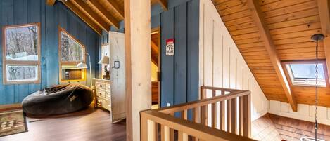 A cozy cabin interior with a loft bedroom, wooden beams, a ceiling fan, a bean bag, and a view of the outdoors.
