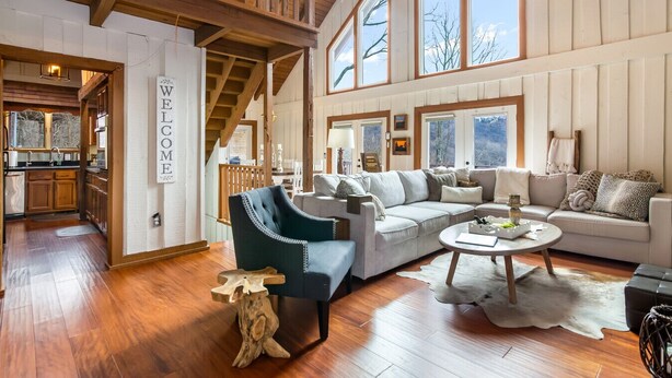 A spacious living room with a high ceiling, wooden beams, and large windows offering a view of the outdoors. - King Bed, Arcade, Hot Tub, Amazing Views (Jasper)