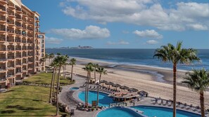 Un hotel con múltiples balcones con vistas a una playa y un área de piscina con sillas de descanso y palmeras.