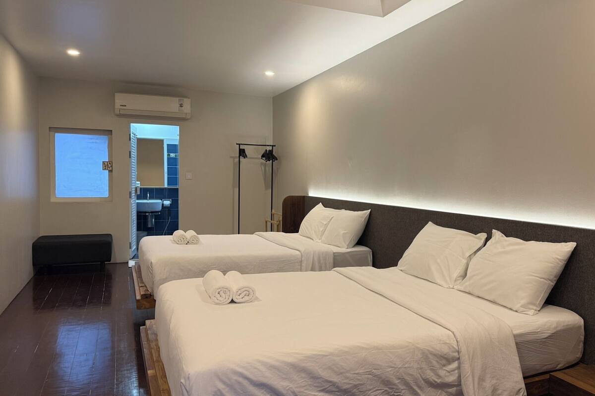 A hotel room with two beds, a bench, a large mirror, and a window.