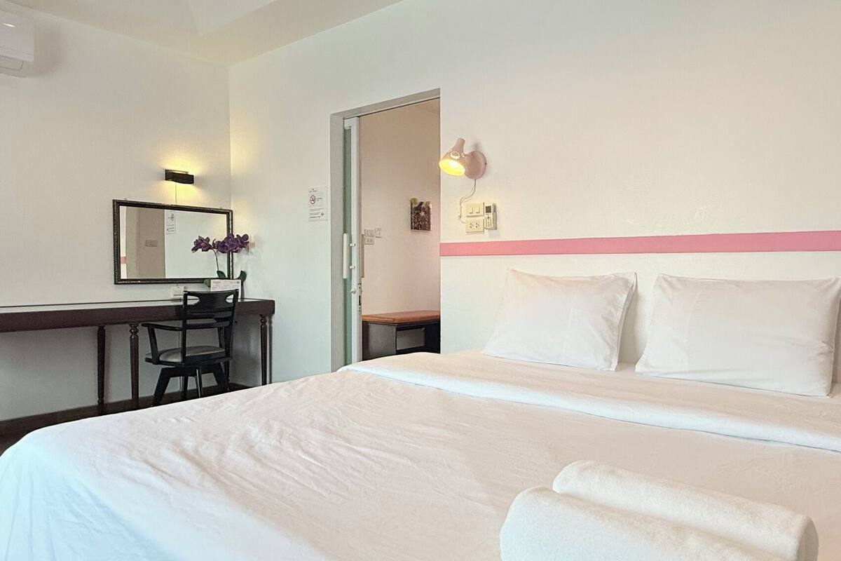A hotel room with a bed, a desk, a chair, a mirror, and a wall-mounted light fixture.