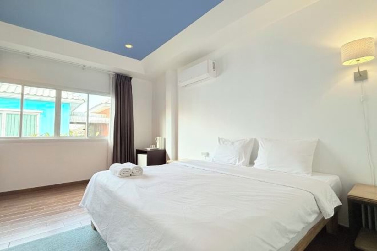 A hotel room with a large bed, a nightstand, a lamp, and a view of a blue building through the window.