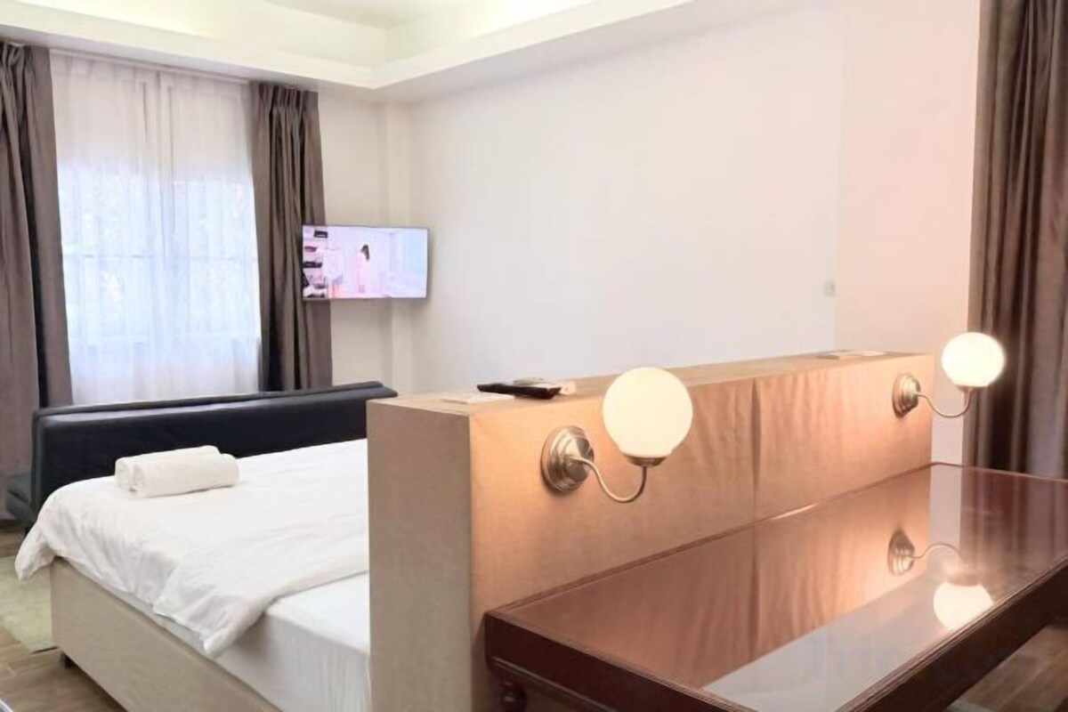 A hotel room with a bed, a wooden headboard, a TV mounted on the wall, and a mirror with lights.