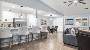 A modern kitchen with white bar stools, a dining area with a gray sofa, and a ceiling fan.