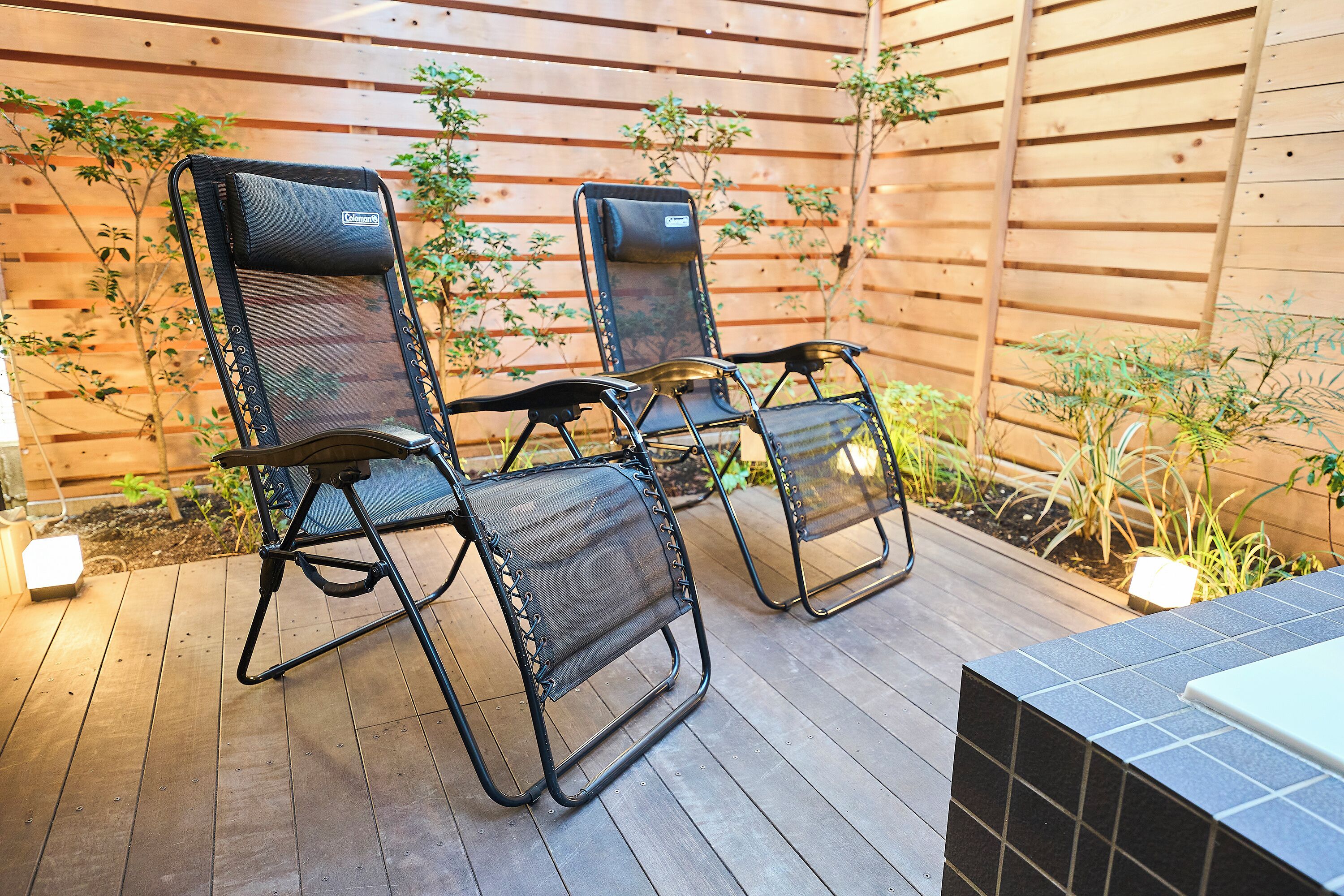 Dos sillas de exterior con respaldo y reposabrazos de malla, ubicadas en una terraza de madera con un cerco de madera y plantas al fondo.