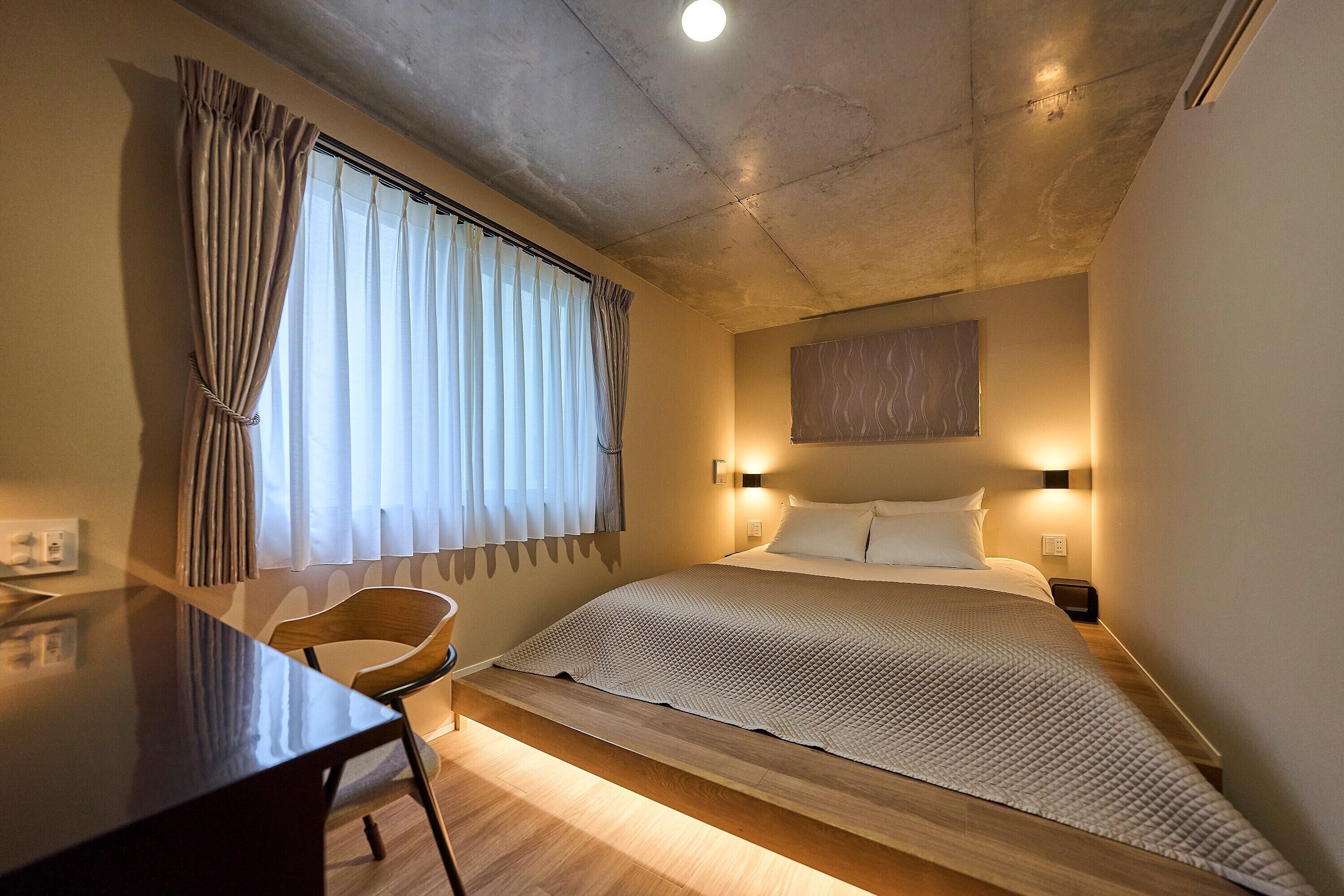 Un dormitorio con cama, mesita de noche, silla y ventana con cortinas.
