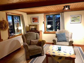 Un salon chaleureux avec deux fauteuils, une table recouverte d’une nappe, un radiateur, un téléviseur et des œuvres d’art accrochées aux murs.