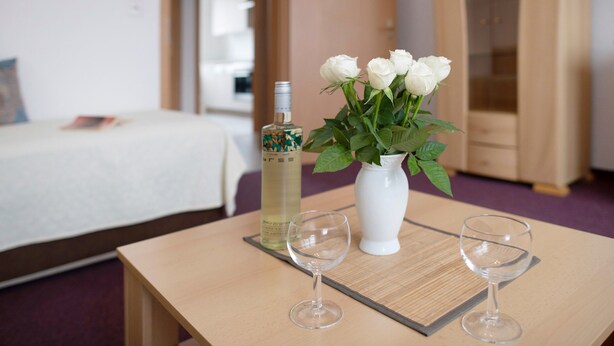 Una camera d'albergo con un letto, un tavolo con due bicchieri e una bottiglia, e un vaso con fiori bianchi.
