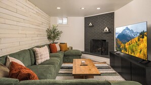 A modern living room with a green sofa, a wooden coffee table, a fireplace, and a large TV on a black stand. - Modern Oasis – 6BR Arvada Retreat, Sleeps 14, Hot Tub, Walkable to Olde Town (Arvada)
