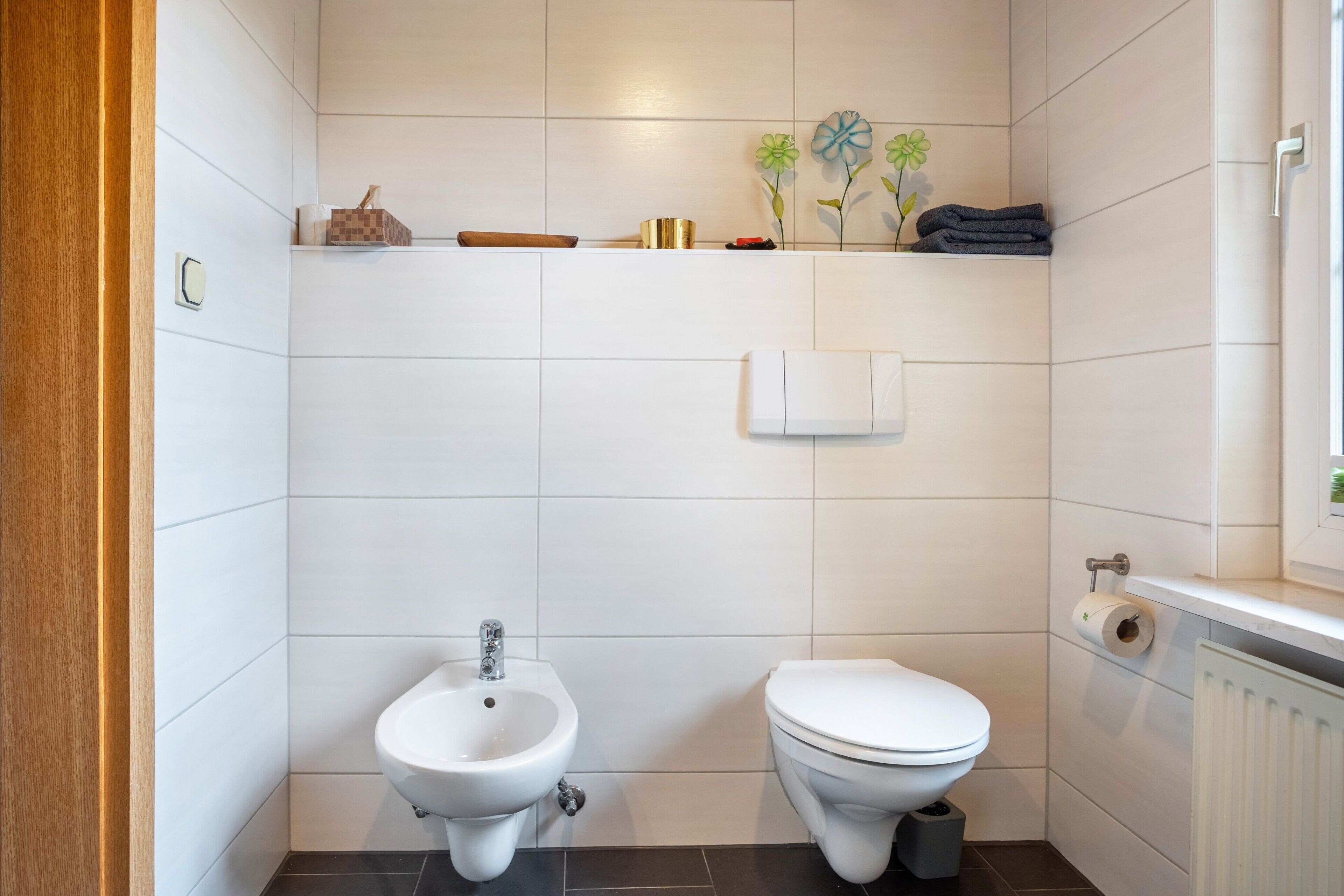A bathroom with white tiles, a bidet, a toilet, a shelf with toiletries, and a radiator.