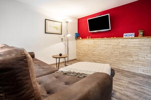 A living room with a sofa, a small table with a lamp, a TV mounted on a red wall, and a stone wall. - Apartment 'Kropp' with Private Terrace, Private Garden and Wi-Fi (Knorrendorf)