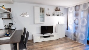Una sala moderna con un televisor de pantalla plana, muebles blancos, una zona de comedor con una mesa y sillas de madera, y un modelo decorativo de barco en la pared.