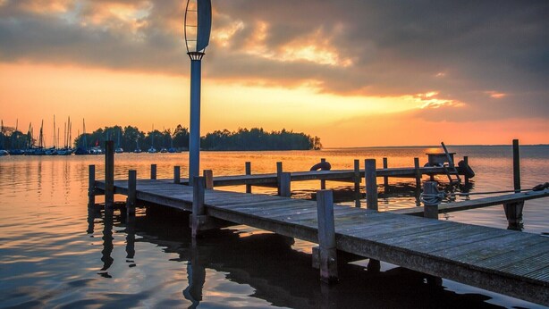 Un muelle de madera se extiende sobre aguas tranquilas, un farol y una orilla lejana con árboles.