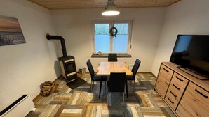 Ein Zimmer mit Holzboden im Fischgrätmuster, einem Esstisch mit Stühlen, einem Fernseher auf einem Holzschrank, einem Kamin und einem Fenster mit Jalousien.