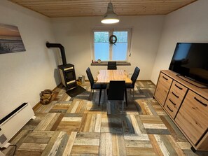 Huoneessa on puulattia, jossa on kalanruotokuvio, ruokapöytä tuoleineen, televisio puisen kaapin päällä, takka ja ikkuna, jossa on sälekaihtimet.