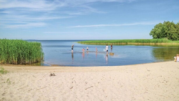 Ein Strand mit Personen, die im flachen Wasser waten, einem sandigen Ufer und einer Reihe hoher Gräser.