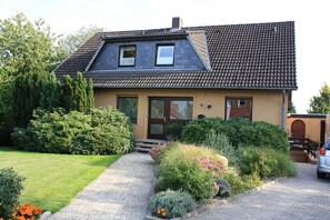 Une maison à deux étages, avec un toit recouvert de tuiles, une porte d’entrée et un petit jardin devant, avec un chemin menant à l’entrée.