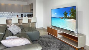 A modern living room with a large flat-screen TV, a grey sofa with patterned cushions, a kitchen area with white cabinets and a brick backsplash, and a black and white patterned rug.