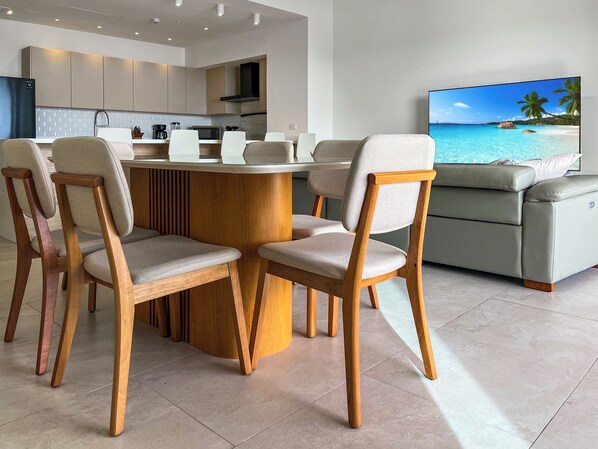 A modern kitchen with built-in appliances, a dining area with wooden chairs and a round table, and a living area with a sofa and a TV displaying a beach scene.