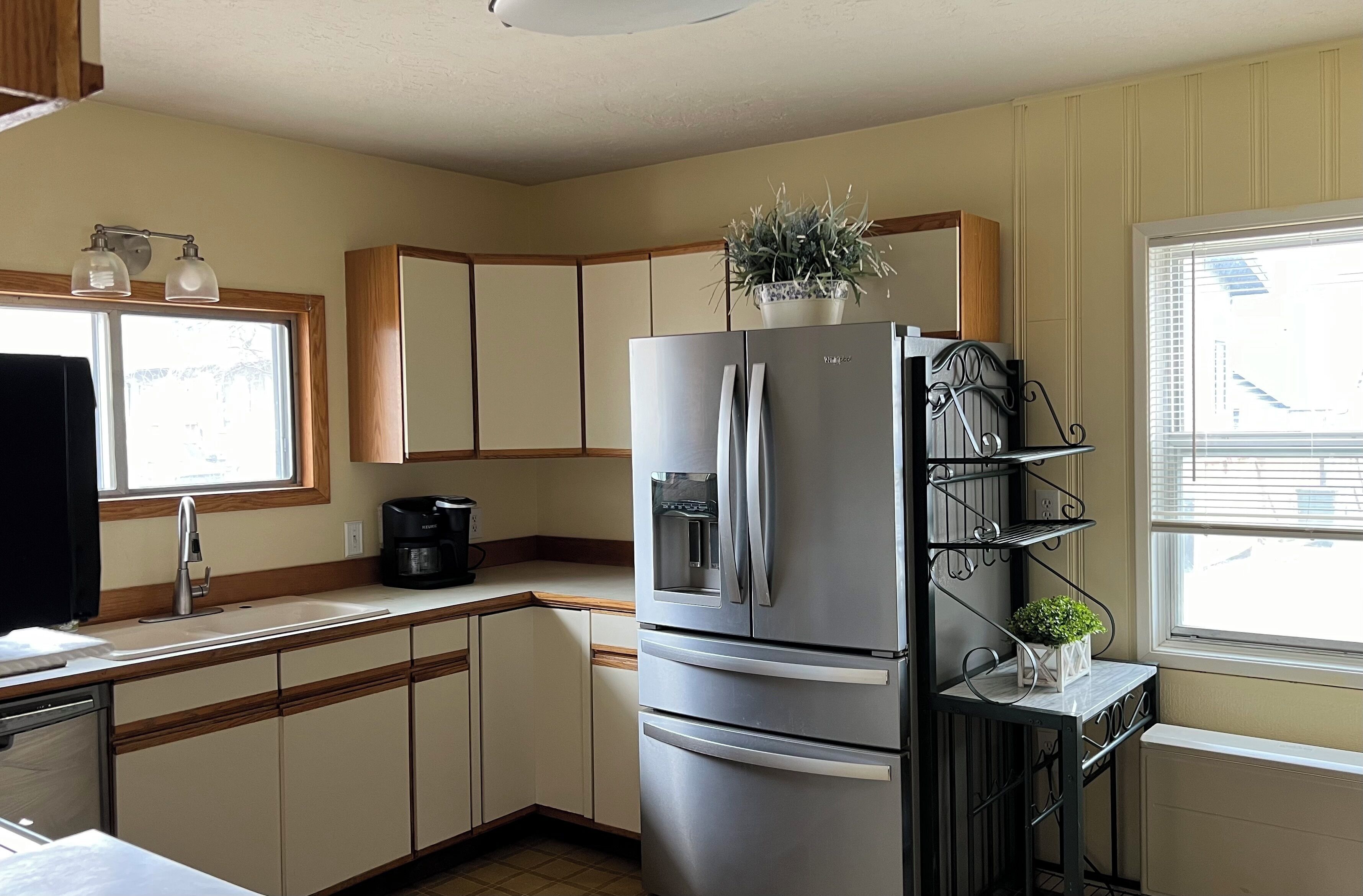 A kitchen with a stainless steel refrigerator, wooden cabinets, a window with blinds, and a small shelf with a plant.