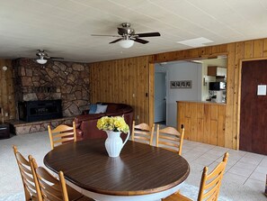 A dining area with a round wooden table, four chairs, and a vase of flowers. There is a stone fireplace and a ceiling fan above the table.