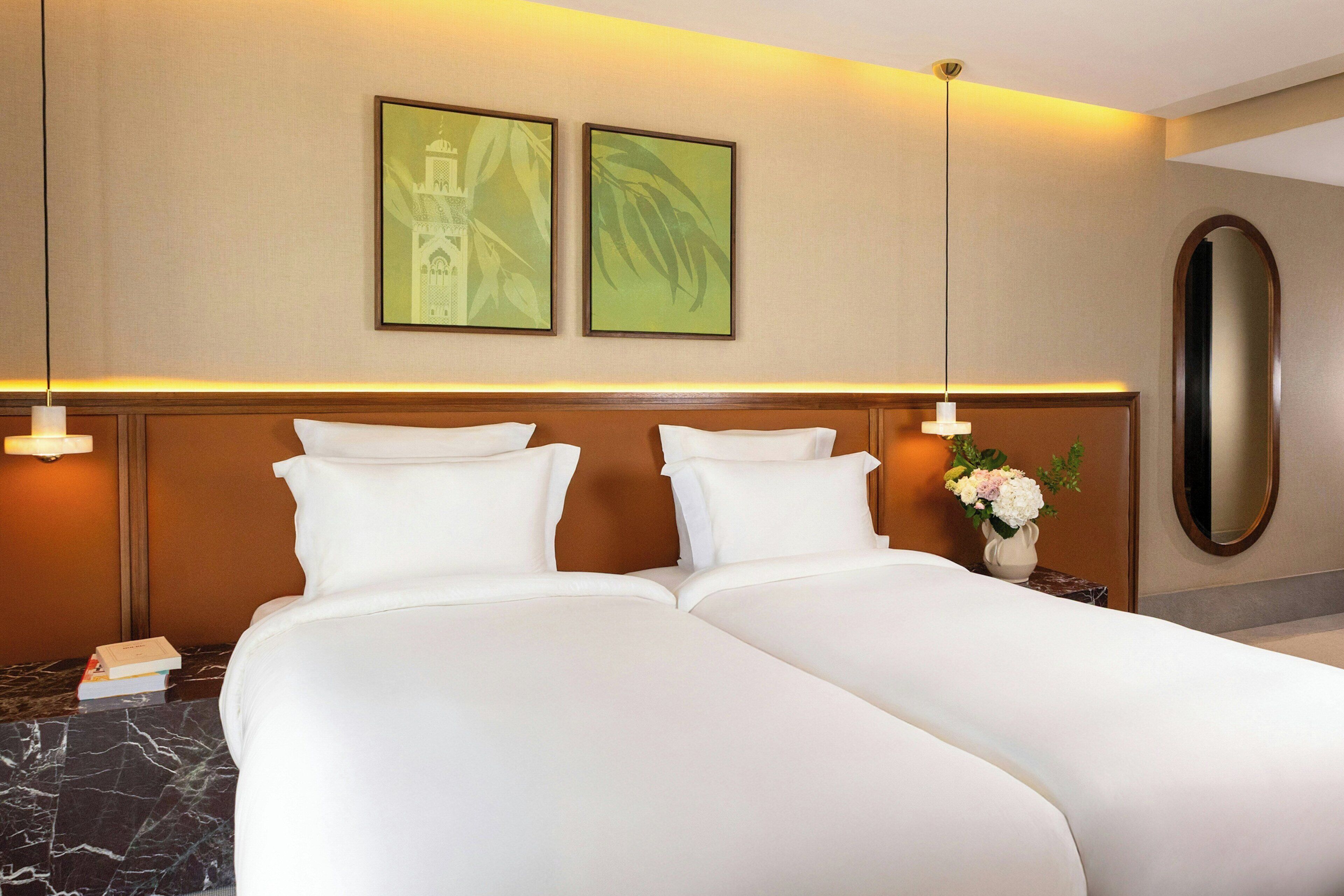 A hotel room with two beds, a large mirror, a nightstand with a vase of flowers, and two framed pictures on the wall.