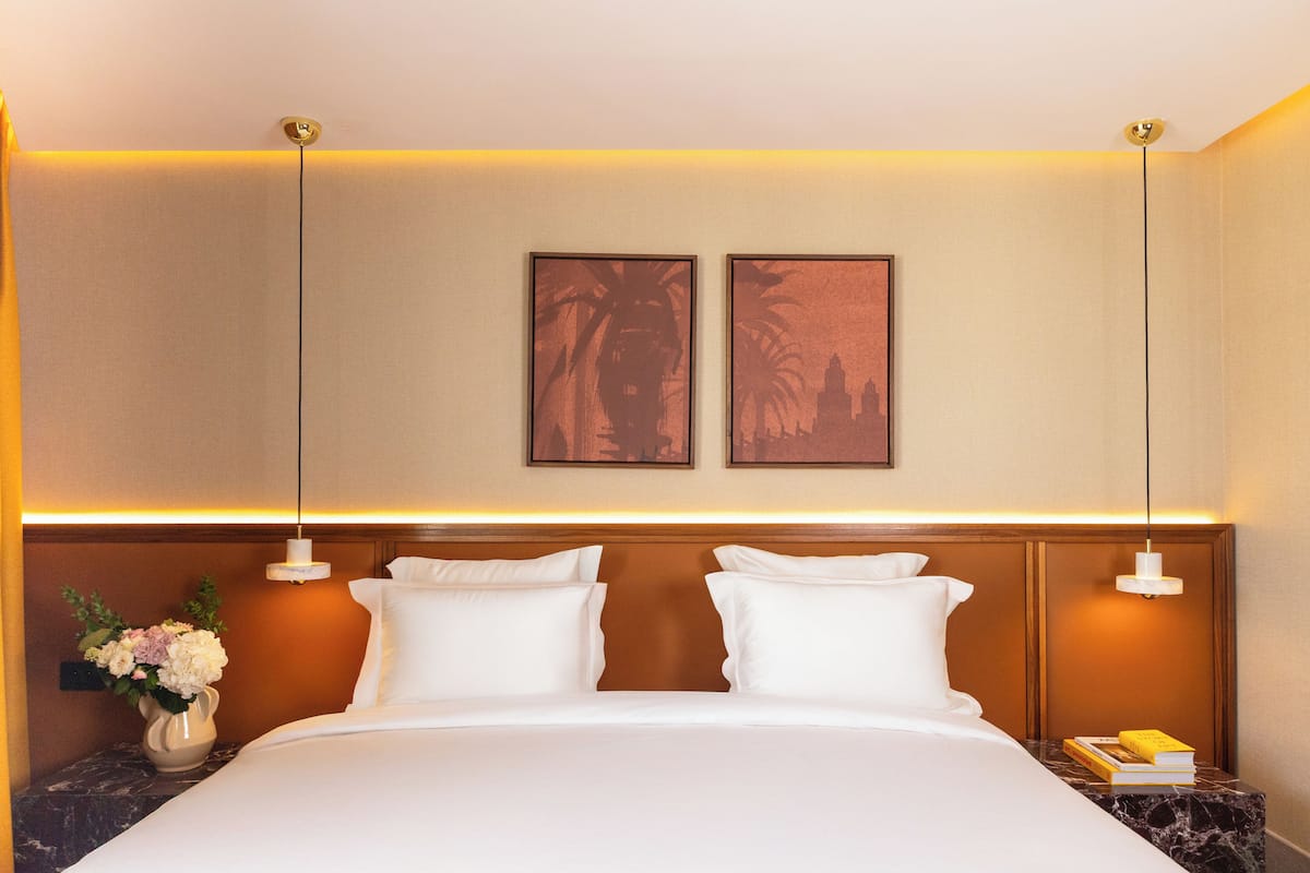 Habitación de hotel con una cama grande, dos marcos de cuadros en la pared, una mesita de noche con un jarrón de flores y un libro.