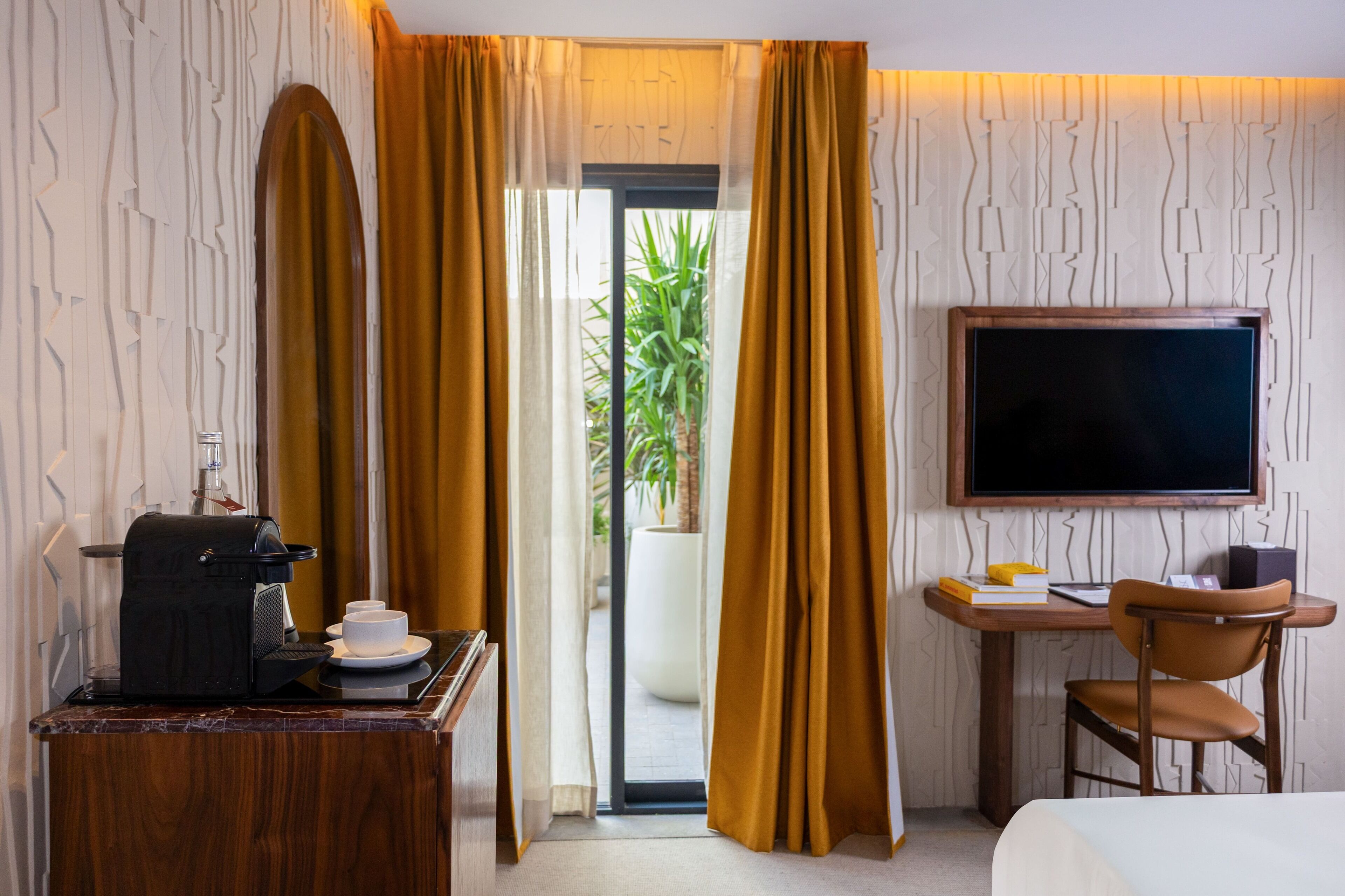 Habitación de hotel con escritorio, silla, televisor, cafetera y vista a zonas verdes.