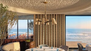 Ein Zimmer mit rundem Tisch, Stühlen, Blick auf den Strand und einem gemusterten Vorhang.