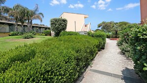 A pathway flanked by neatly trimmed hedges and a building with a flat roof and a staircase.
