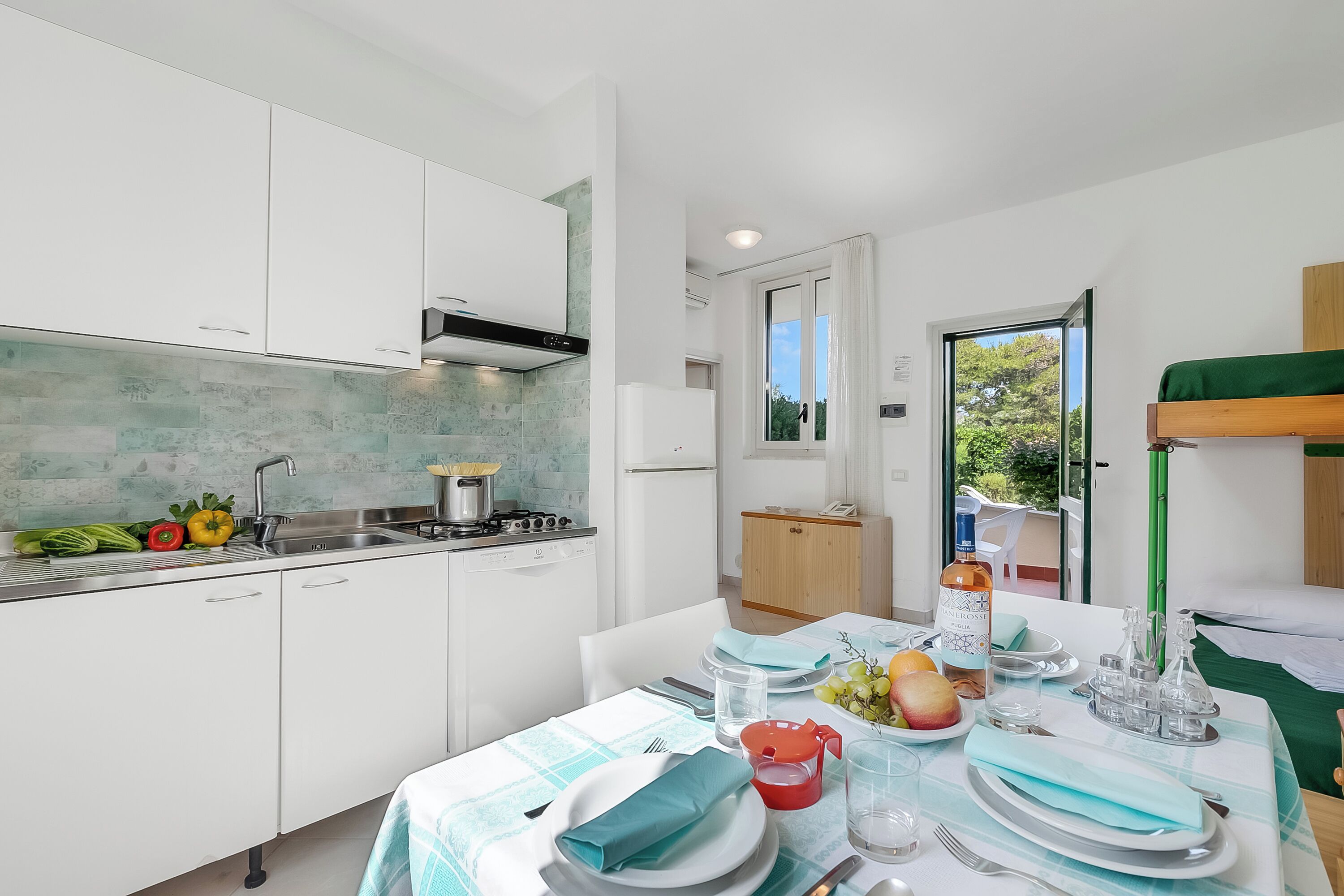 Une cuisine moderne avec des meubles blancs, une cuisinière et un évier. On y trouve un coin repas avec une table dressée pour deux personnes, une fenêtre donnant sur un espace verdoyant et une porte menant à une autre pièce.