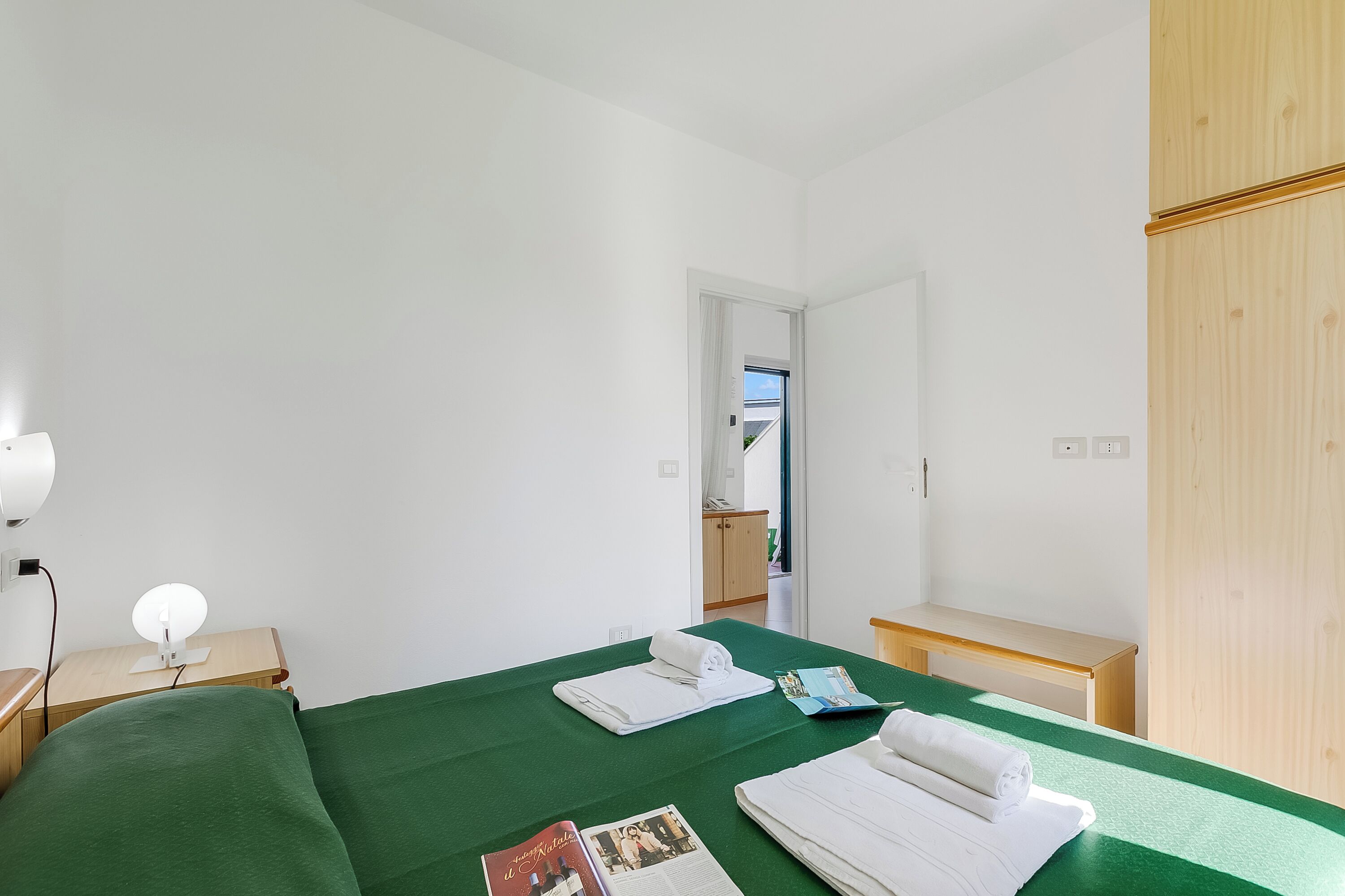 A hotel room with a green bedspread, a bedside table with a lamp, a wooden wardrobe, and a view of the outside through an open door.