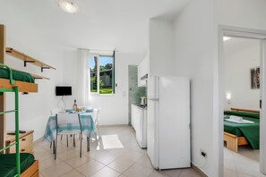 Ein kompaktes Zimmer mit Etagenbett, Essbereich inklusive Tisch und Stühlen, Küche mit Kühlschrank und einem Bett mit grüner Bettwäsche.