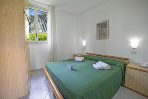 A hotel room with a bed, a bedside table, a window with curtains, a wall-mounted light, and a framed picture.