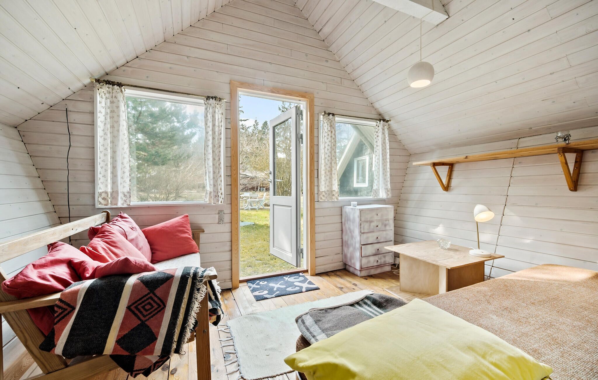 Une chambre mansardée chaleureuse avec un plafond en pente, des murs en bois, un lit agrémenté de coussins rouges et jaunes, une petite table avec une lampe et une fenêtre donnant sur un jardin.