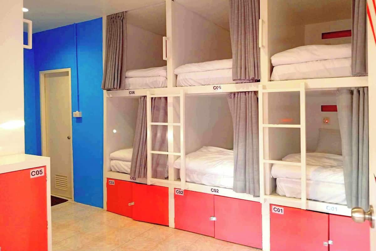 Quarto de dormitório com beliches, cada um identificado com a letra C seguida de um número. Existem armários vermelhos sob as camas, e uma porta no lado esquerdo.
