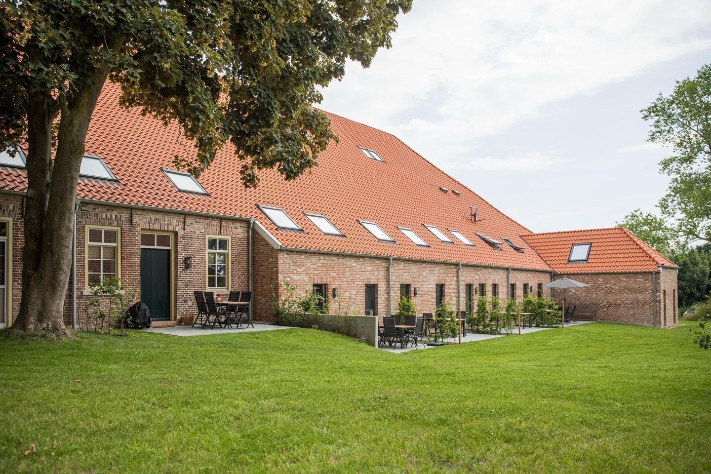 Un bâtiment en briques avec un toit à pente recouvert de tuiles rouges, de nombreuses fenêtres de toit et une cour verdoyante aménagée avec des sièges extérieurs.