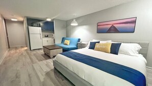 A bedroom with a bed, bedside tables, a nightstand, a desk, a chair, and a kitchenette area.