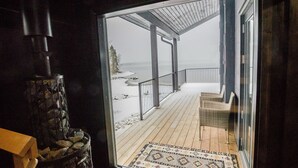 A wooden deck with a patterned rug, a stone fireplace, and a view of a snowy beach through a glass door. - Lakeside villa by Interhome (Nurmes)