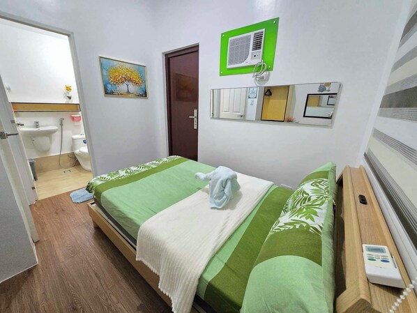 Una habitación de hotel con cama, mesita de noche, unidad de aire acondicionado en la pared, un cuadro, una puerta y un baño visible a través de una puerta abierta.