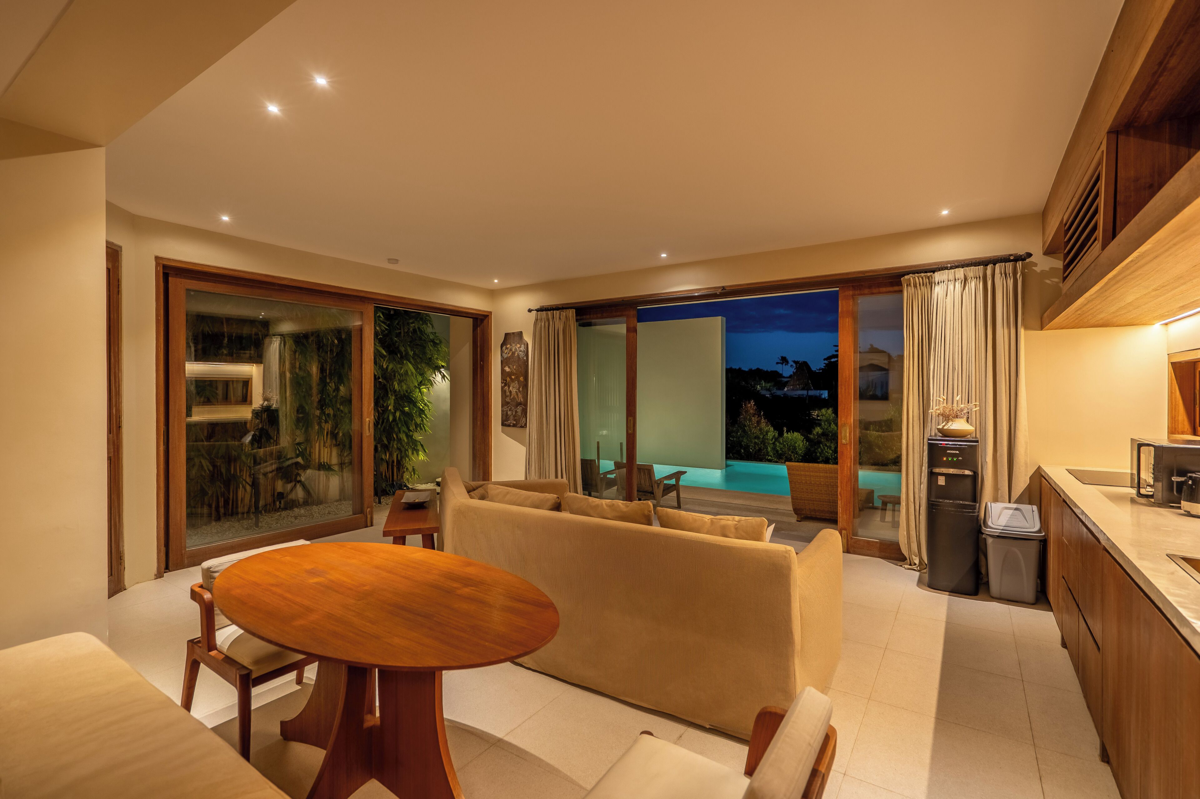 A modern living room with a sofa, a wooden table, and a kitchen area with a refrigerator and sink.