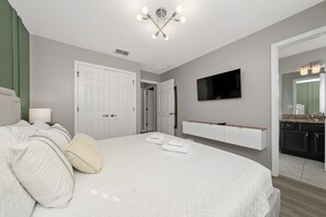 A bedroom with a large bed, a TV mounted on the wall, and a bathroom visible through an open door. - Stay Close to Magic 3BD No Carpet Home w/ Pool (Kissimmee)