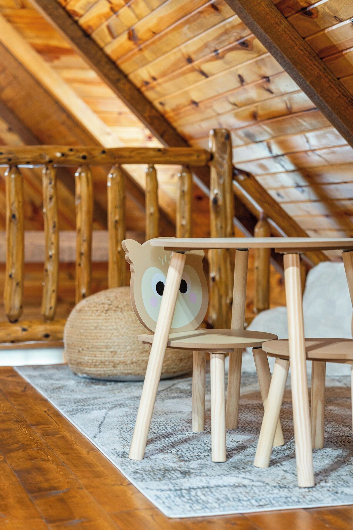 Una silla de madera con diseño de cara de búho recortada, una cesta tejida y una mesa de madera en una habitación con techo inclinado y vigas de madera.