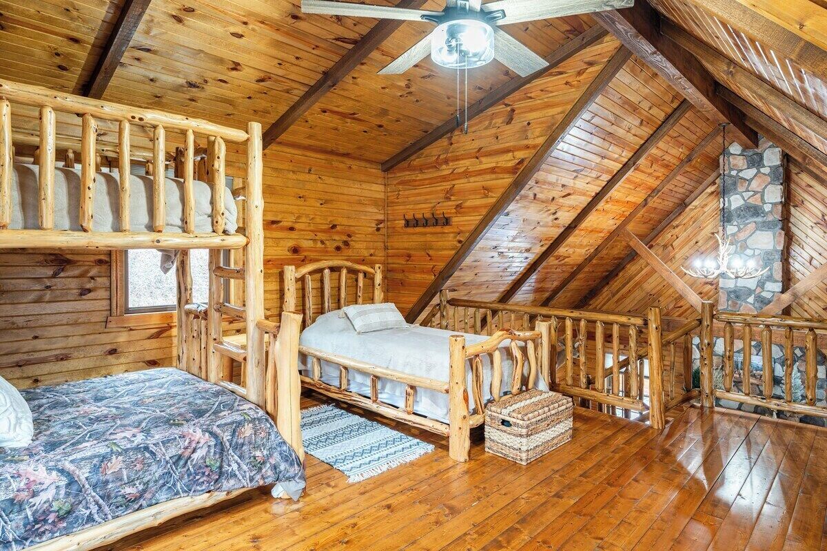 A wooden cabin with a loft bedroom, a bunk bed, a single bed, and a stone fireplace.
