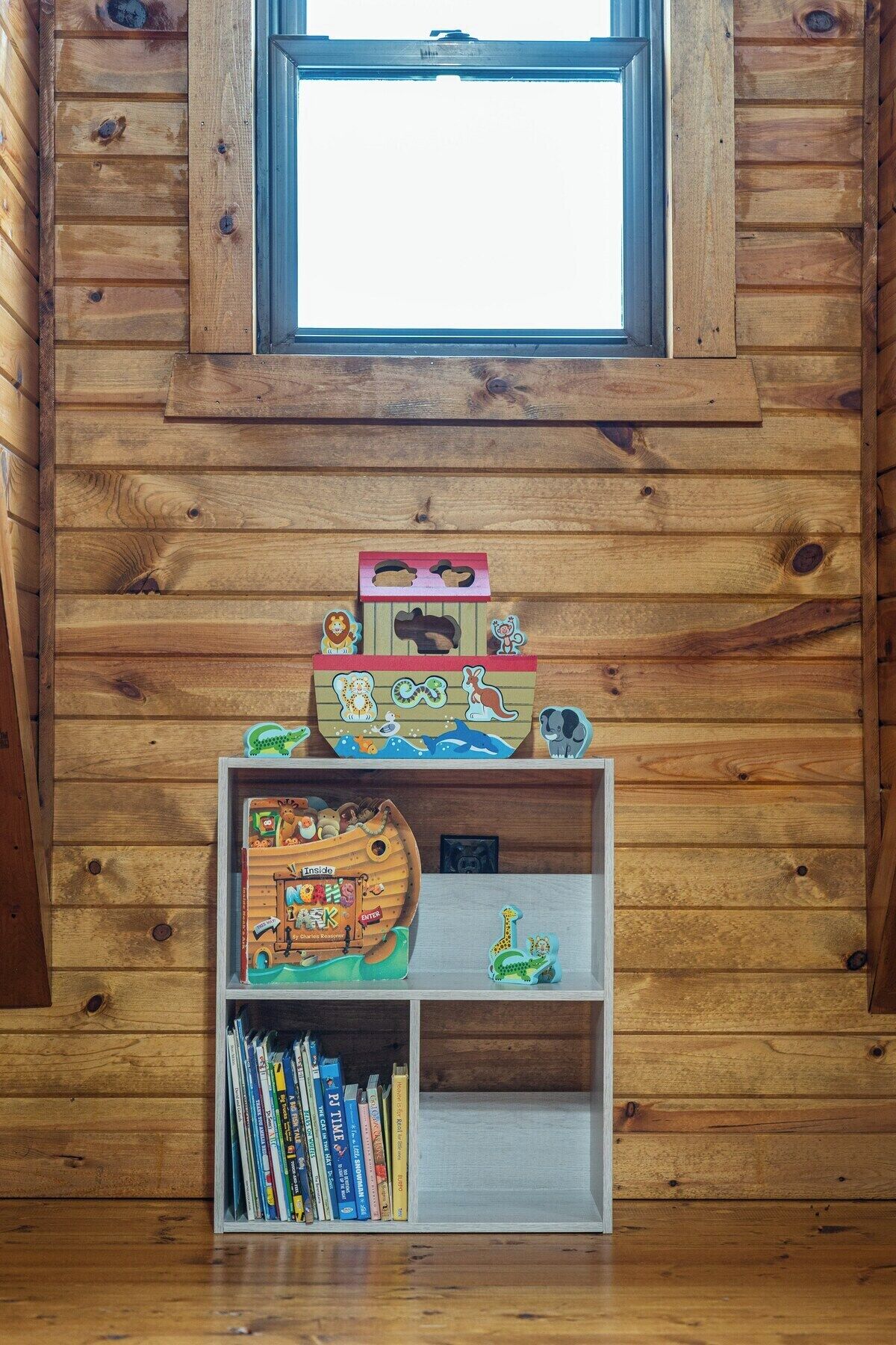 Interior de una cabaña de madera con una estantería llena de libros infantiles y un barco de juguete.