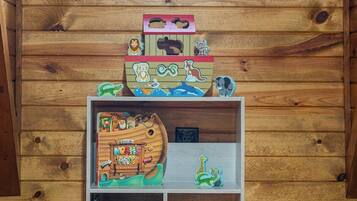 Interior de una cabaña de madera con una estantería llena de libros infantiles y un barco de juguete.
