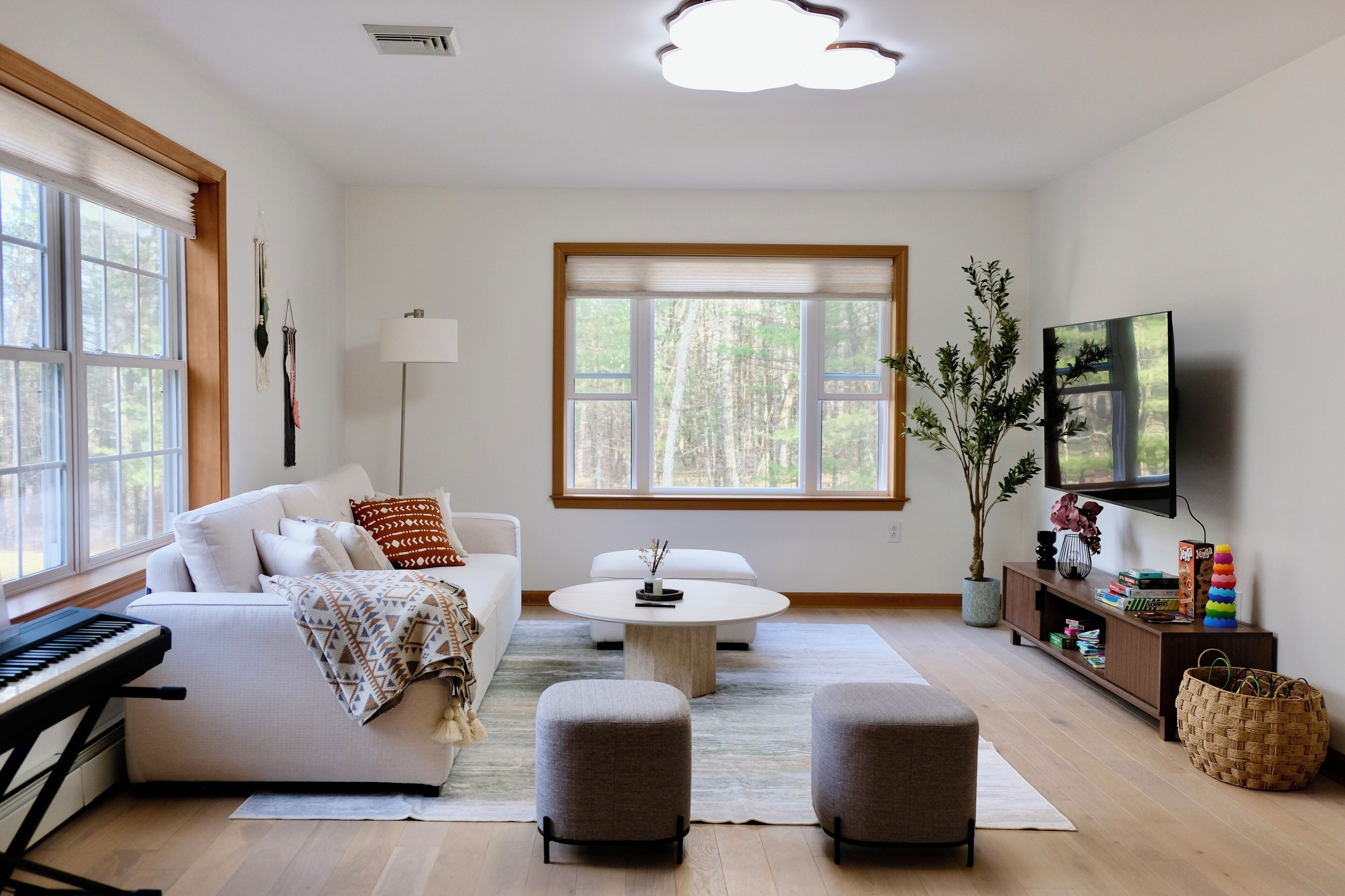 A modern living room with a white sofa, a round coffee table, a keyboard, a TV, and a large window with a view of trees.