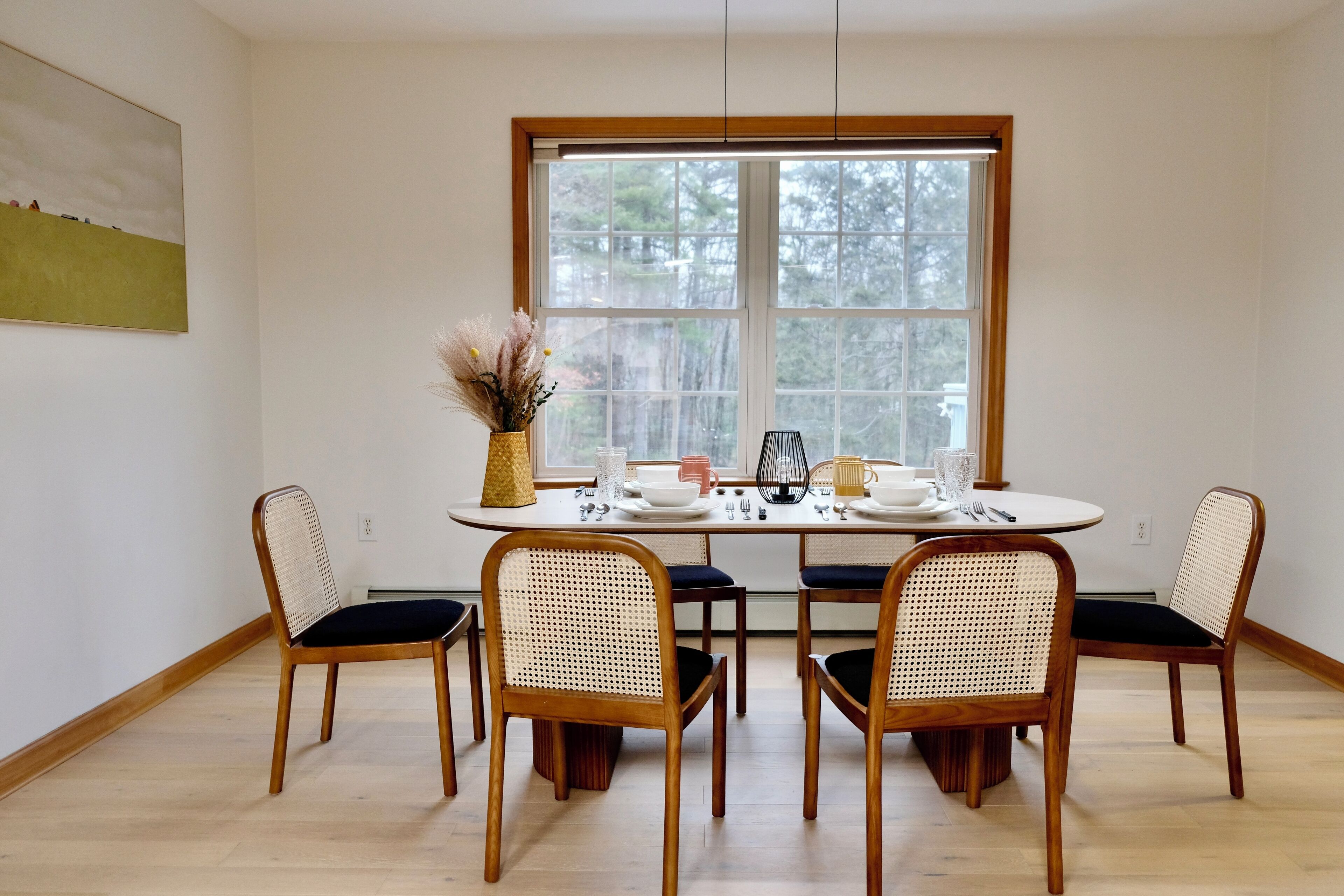 A dining area with a round table set for four, surrounded by wooden chairs with woven backs. A window with a view of trees is behind the table.