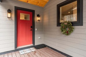 A red door with a welcome mat, two wall-mounted lanterns, and a wreath on the wall.