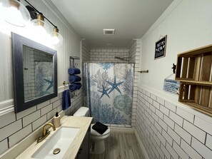 A bathroom with a white tiled wall, a shower curtain with a starfish and coral pattern, a toilet, a sink with a gold faucet, and a mirror with a black frame. - Pelican Place--lit dock, crab cages, W/D, 5 TVs, gameroom, fully stocked kitchen (Palacios)